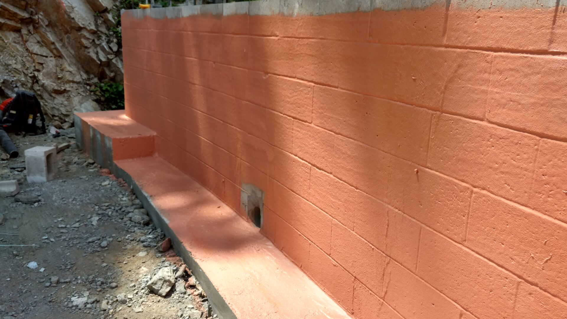 A newly constructed retaining wall made of concrete blocks with an orange coating, featuring a small drain hole near the base. Construction debris is scattered on the ground nearby.